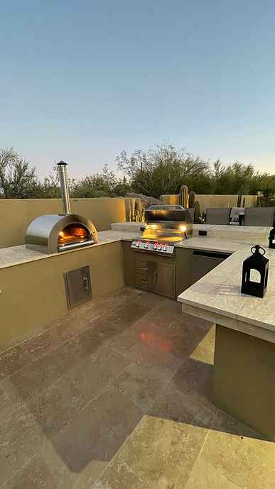 Outdoor kitchen setup with a stainless steel wood-fired pizza oven, built-in gas grill, stone countertops, and a tiled patio under a clear evening sky.