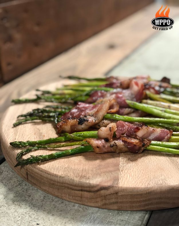 Grilled asparagus spears wrapped with bacon on a wooden cutting board, with WWPO logo in the corner.